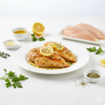 A slightly angled, close-but-not-too-close composition of Baked Lemon Chicken surrounded by its raw ingredients on a bright white kitchen surface. Include small bowls or dishes containing olive oil, minced garlic, dried oregano, thyme, lemon halves, and fresh parsley alongside raw chicken breasts. The finished baked chicken dish is placed prominently in the center on a white plate, with visible lemon slices and herb garnish. The setup is clean, balanced, and realistic, showcasing both preparation and completion. Hyper-real clarity, bright even lighting, pure white background, no shadows, no yellow tones, crisp and modern homemade editorial style.
