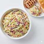 A detailed top-down view of Creamy Coleslaw Homemade photographed from above on a pure bright white surface. The creamy coleslaw is presented in a simple white bowl, showing the colorful mix of green and red cabbage with shredded carrots coated in a smooth, glossy dressing. A few small portions of coleslaw are neatly served on a plate beside grilled meat or a sandwich for context. The composition is clean, minimal, and fresh, emphasizing texture and natural color contrast. Hyper-real clarity, pure bright white background, neutral cool lighting, no shadows, no warm tones, crisp and modern homemade presentation.